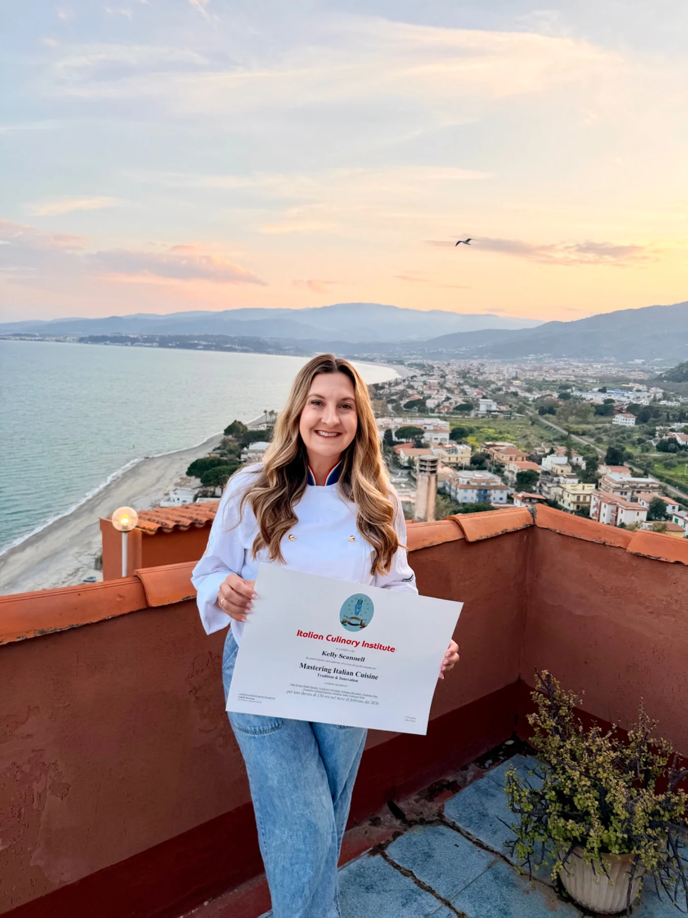 Chef Kelly Scannell holding her culinary certificate with a coastal view behind her