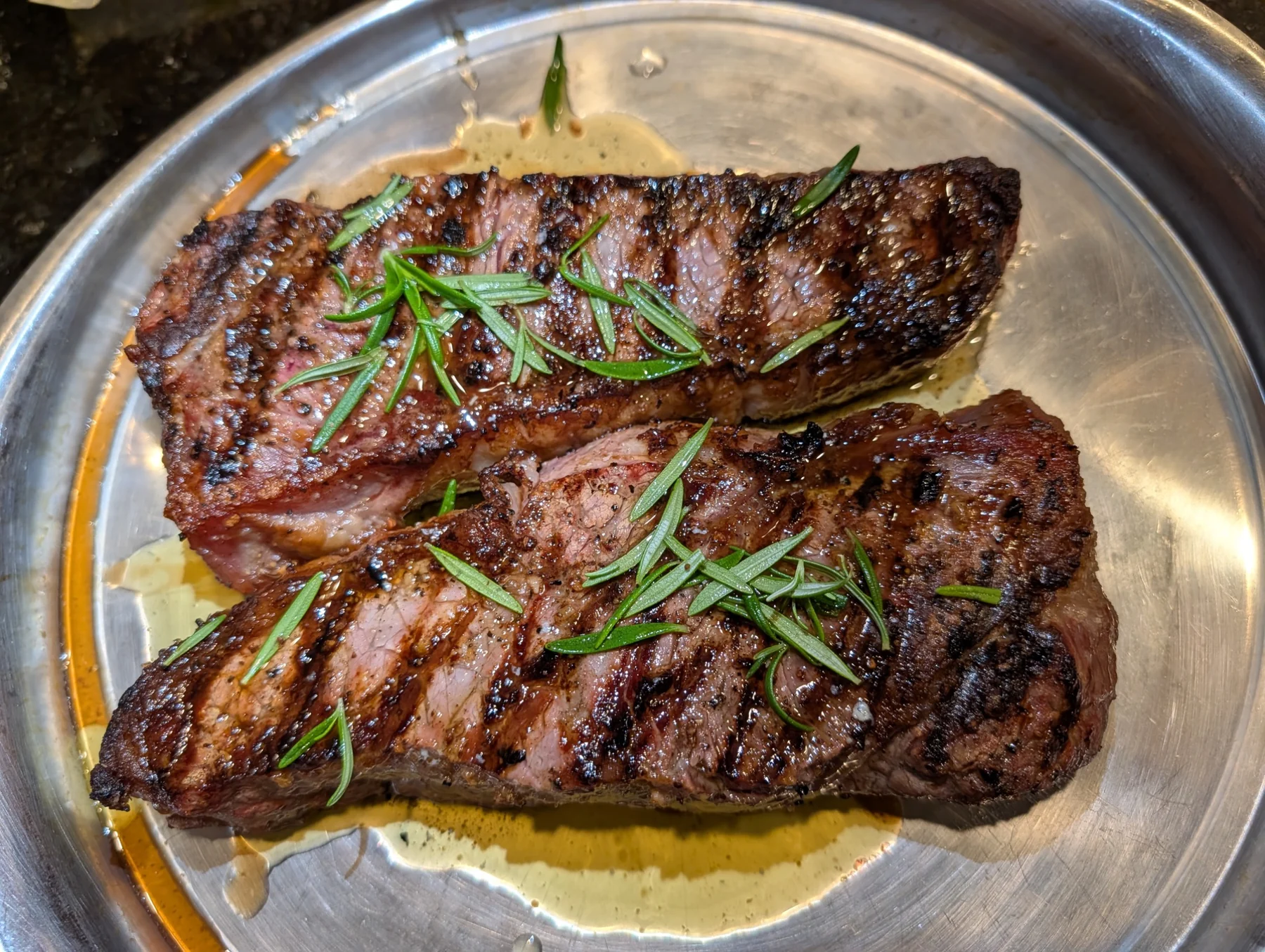 Two seared steaks topped with fresh rosemary on a silver platter