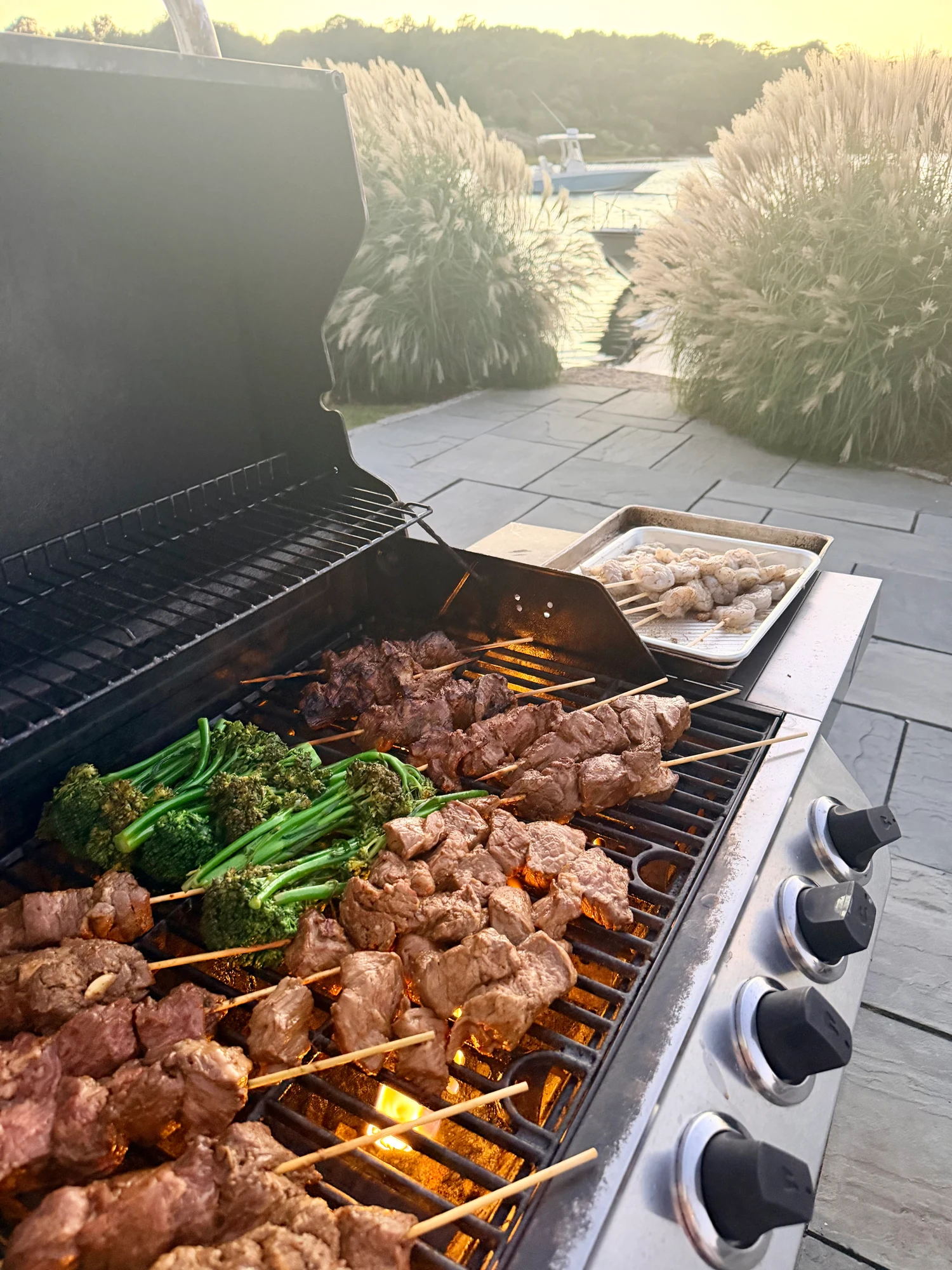 Steak skewers and broccolini on an outdoor grill, with seafood skewers and a coastal harbor view nearby